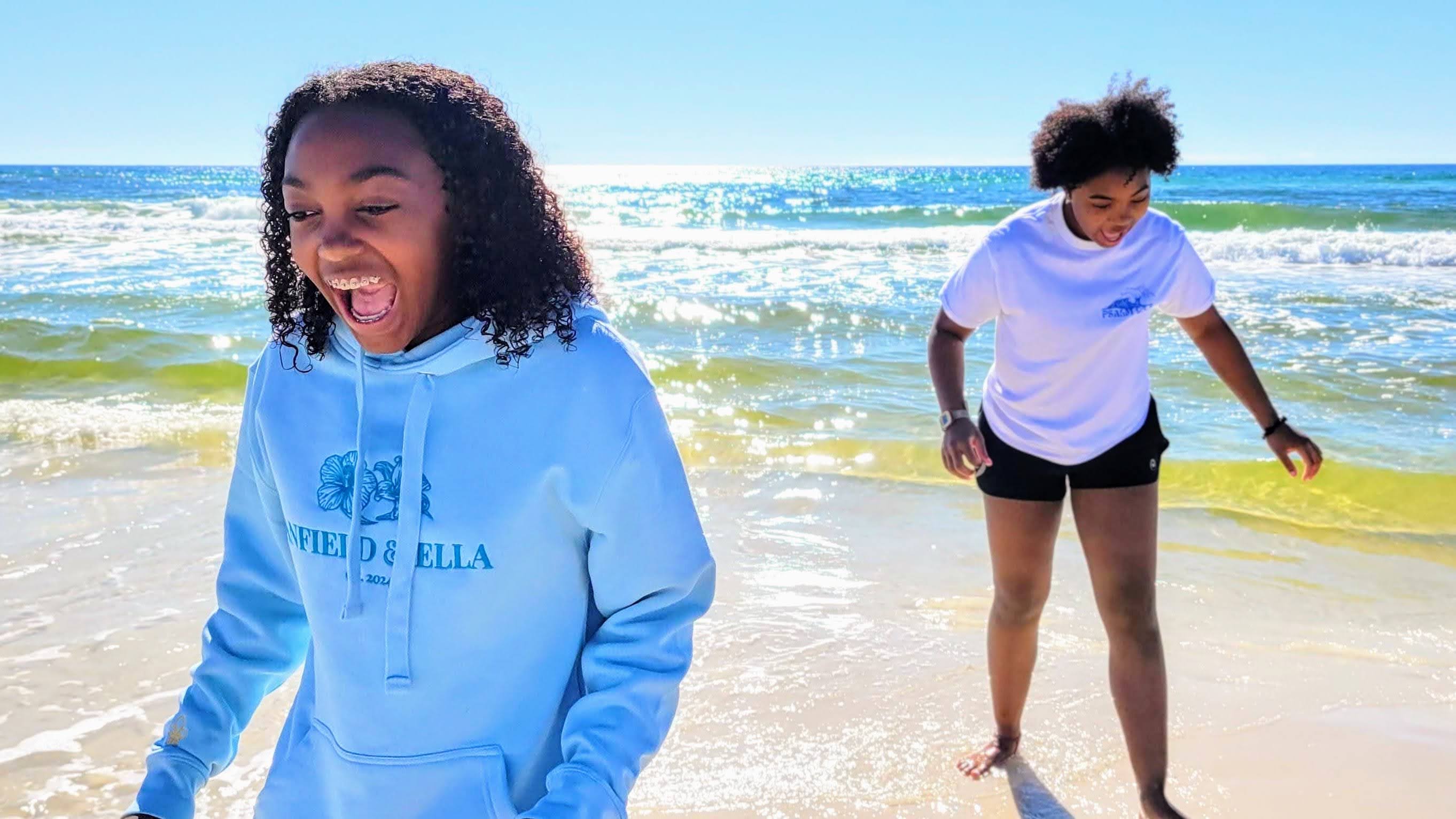 Anfield & Ella | Cover Photo - A teen and tween girl play at the beach wearing the Sky Blue Anfield & Ella Founder's Edition Hoodie and the White Sea Sky Land Tee | Anfield and Ella